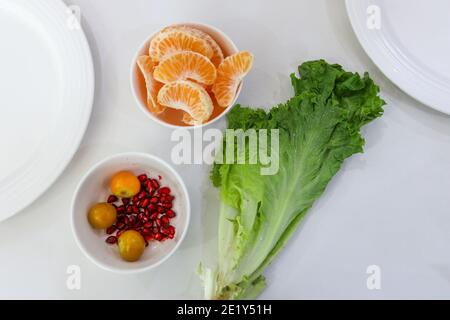 Freshly peeled oranges with pomegranate and celery leaves arranged on a table with partial white plates in corners Stock Photo