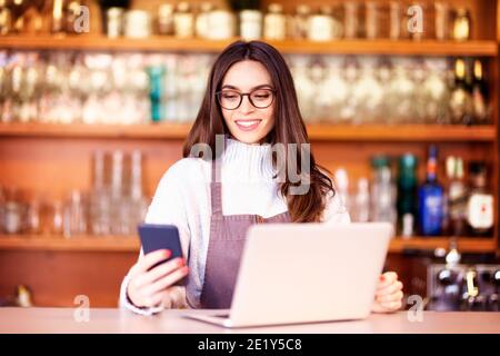 Portrait of a beautiful waitress text messaging and using laptop while standing behind the counter at a cafe. Small business. Stock Photo
