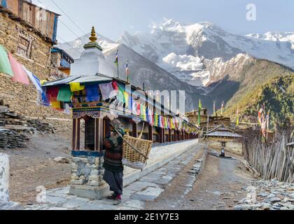 Old local woman spinning prayer wheels along a Mani wall with a snow avalanche on Annapurna II mountain in the background, Upper Pisang, Nepal Stock Photo