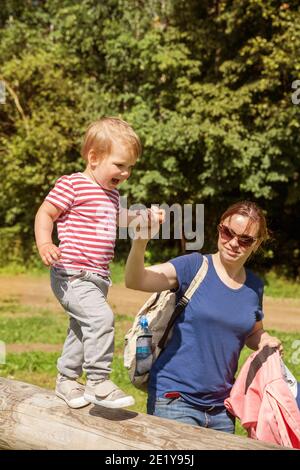 Family on walk in park. Mom helps baby to keep balance on balance beam. The child is happy and delighted Stock Photo