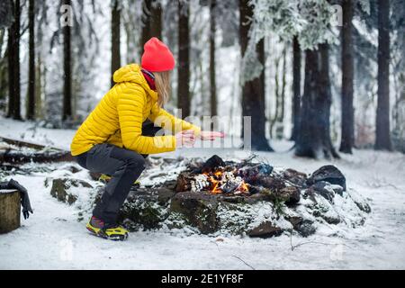 A beautiful blonde tourist in a yellow down jacket drinks tea and warms her hands by the fire. Stock Photo