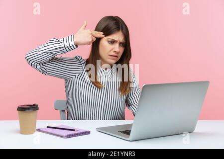 Depressed woman office worker holding finger gun near head talking video call on laptop, expressing extreme fatigue and boredom from job. Indoor studi Stock Photo