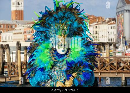 Woman dressed in traditional decorated costume with feathers and painted mask during Venice Carnival with St Mark's Campanile behind Venice Italy Stock Photo