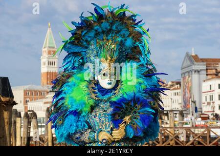 Woman dressed in traditional decorated costume with feathers and painted mask during Venice Carnival with St Mark's Campanile behind Venice Italy Stock Photo