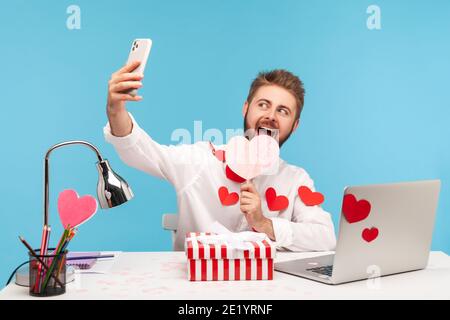 Funny happy man with beard in white shirt with red sticky hearts making selfie with pink heart and striped giftbox sitting at workplace. Indoor studio Stock Photo