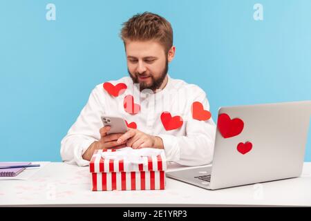 Smiling bearded businessman in white shirt in red heart shaped stickers sitting at desk with laptop and striped giftbox typing on smartphone. Indoor s Stock Photo
