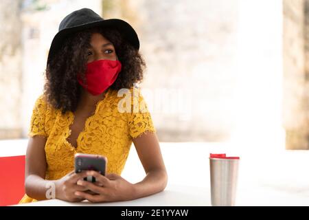 young black woman with curly hair, with red mask, yellow dress and black hat, checking her phone Stock Photo