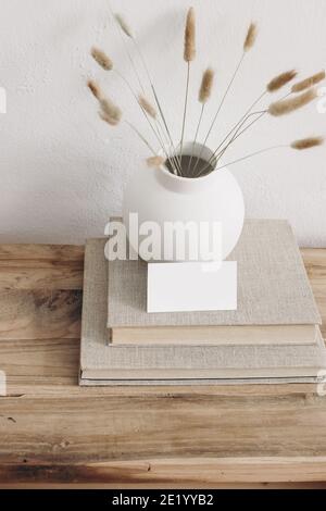 Business card mockup on vintage wooden table. Modern ceramic vase with dry Lagurus ovatus grass and pile of books. White wall background. Scandinavian Stock Photo