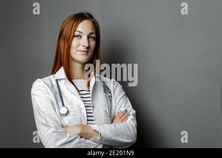 Portrait of glad smiling doctor in white uniform standing with crossed hands on gray background. Stock Photo