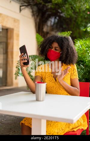 young black woman with curly hair, with red mask, yellow dress and black hat, checking her phone Stock Photo