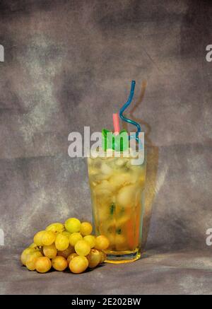 A tall glass of a light drink with ld and a bunch of white grapes nearby, stand on an abstract background. Close-up. Stock Photo