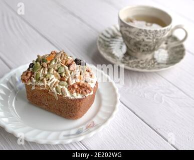 Carrot cake with mixed nuts, seeds and dried fruits as topping with cup of tea at the background. On wood table. Stock Photo