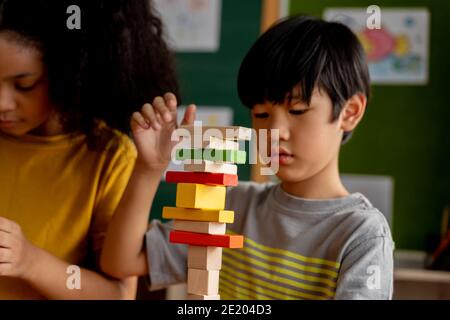 ocused Asian boy building tower with wooden blocks while playing near African American girl in school Stock Photo