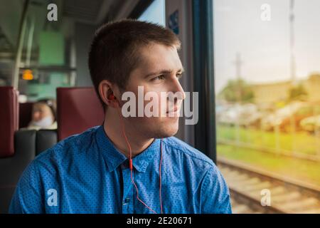 Young european guy in a blue shirt listening to music on headphones looks out the window while traveling on a modern electric train. Stock Photo