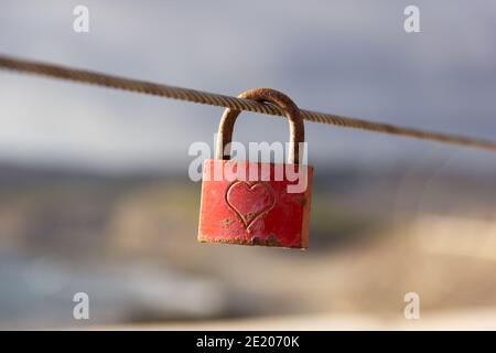 Red love padlock attached to steel wire. Rusty lock with heart shape carved hanging on railing outdoors. Valentines day, unity, memory concepts Stock Photo