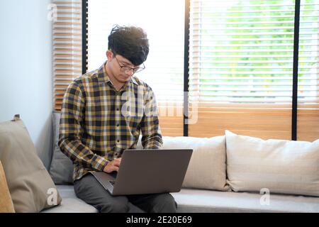 young attractive asian man relaxing comfortably working on laptop in bright living room at his home Stock Photo
