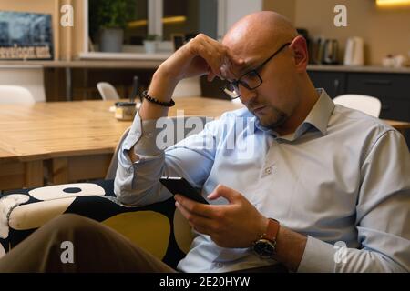 Caucasian mid adult man using smartphone while sitting on couch at home, playing games, reading messages Stock Photo