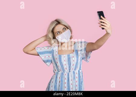 Charming lady is making a selfie on a pink wall while wearing a mask and summer dress Stock Photo