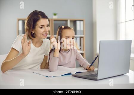 Happy mom and daughter waving hands at laptop, greeting teacher in online lesson Stock Photo