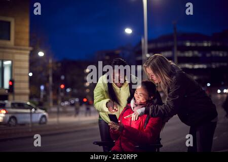 Woman In Wheelchair Having Night Out With Friends In City Ordering Taxi Using Mobile Phone App Stock Photo