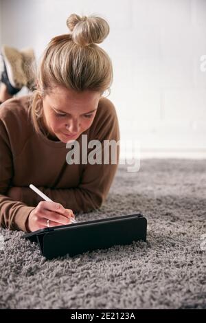 Woman Drawing On Digital Tablet Using Stylus Pen Lying On Carpet At Home Stock Photo