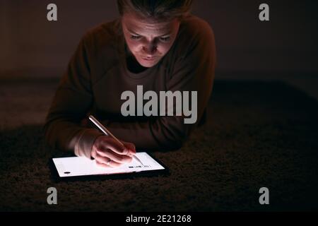 Close Up Of Woman Drawing On Digital Tablet Using Stylus Pen Lying On Carpet At Night Stock Photo