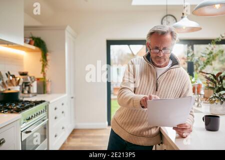 Mature Man At Home In Kitchen Drinking Coffee And Looking At Financial Documents Stock Photo