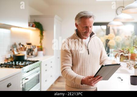 Mature Man In Kitchen Working From Home Using Digital Tablet Stock Photo