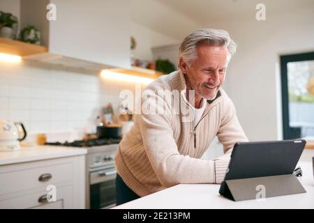 Mature Man In Kitchen Working From Home Using Digital Tablet Stock Photo