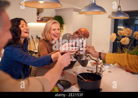 Group Of Mature Friends Making A Toast As They Meet At Home For Meal And Serve Food In Kitchen Stock Photo