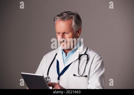 Studio Portrait Of Mature Male Doctor In White Coat Using Digital Tablet Against Grey Background Stock Photo