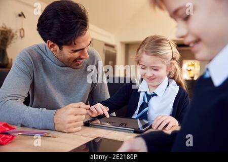 Father Helping Children Using Digital Tablets Wearing School Uniform With Homework At Table Stock Photo