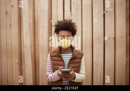 Waist up portrait of teenage African-American boy wearing mask and looking at camera while standing against wooden boards in background, copy space Stock Photo