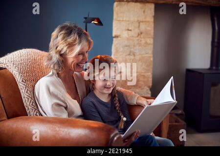 Granddaughter At Home With Grandmother Sitting In Chair Reading Book Together Stock Photo