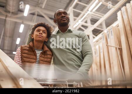 Low angle portrait of African-American father and son shopping together in hardware store while pushing cart with wooden boards for construction or ho Stock Photo