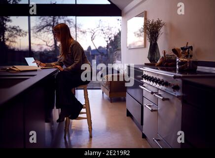 Evening Shot Of Woman In Kitchen Working Or Studying From Home Using Digital Tablet Stock Photo