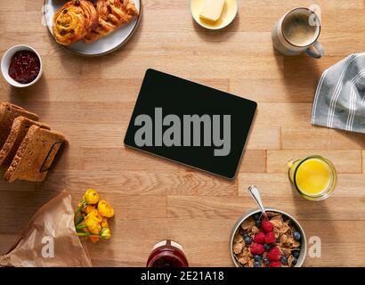 Overhead Flat Lay Of Digital Tablet On Table Laid For Breakfast With Cereal Croissant And Flowers Stock Photo