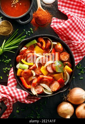Stewed pepper and sausage with tomatoes in a cast iron skillet on a black background, top view. Traditional Hungarian stew called lecho Stock Photo