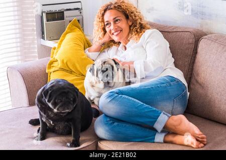 Portrait of cheerful young adult beautiful woman with two dogs pug sitting and relaxing on the sofa at home - concept of animal owner and daily real l Stock Photo