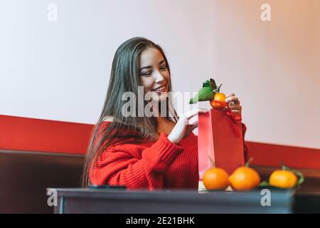 Beautiful smiling young asian woman in red clothes with gift bag with mandarins in cafe, celebrating Chinese New Year Stock Photo