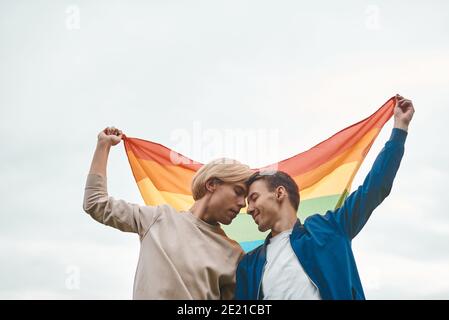 Romantic gay couple hugging, kissing and holding hands outdoors. Two handsome men holding LGBT pride flag. Stock Photo