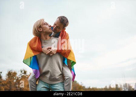 Romantic gay couple hugging, kissing and holding hands outdoors. Two handsome men holding LGBT pride flag. Stock Photo