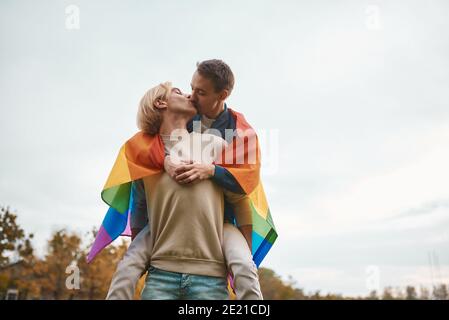 Romantic gay couple hugging, kissing and holding hands outdoors. Two handsome men holding LGBT pride flag. Stock Photo
