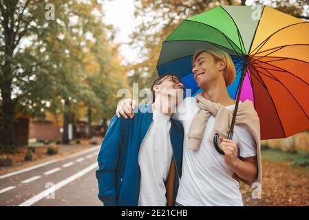 Loving gay couple walking outdoors with rainbow umbrella. Two handsome men having romantic date in park. LGBT concept. Stock Photo