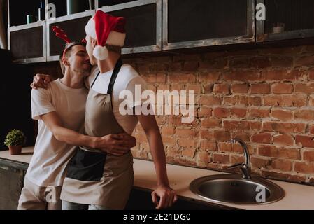 Loving gay couple at home. Two handsome men hugging and kissing on kitchen in Santa Claus hat. Christmas mood. LGBT concept. Stock Photo