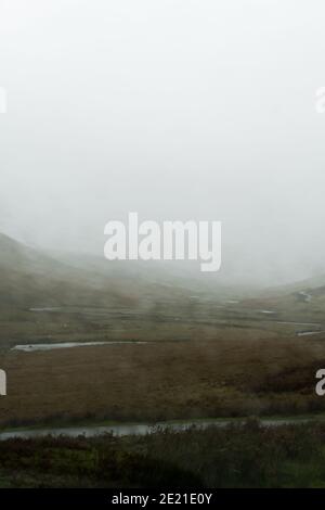 Looking through a car window blurred by raindrops. As a river meanders through a bleak moorland valley. On a moody, misty day. Wales, UK. Stock Photo