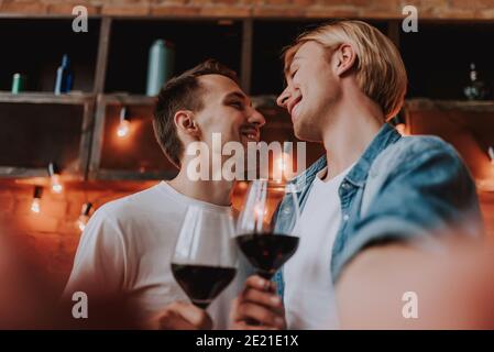 Loving gay couple at home. Two handsome men hugging, kissing and drinking wine on kitchen. LGBT concept. Stock Photo