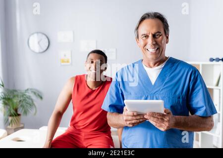 cheerful doctor holding digital tablet near happy african american patient in sportswear on blurred background Stock Photo