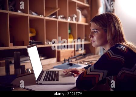 Female Business Owner Working Late In Carpentry Workshop Using Laptop Stock Photo