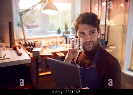 Male Jeweller Making Phone Call Looking At Design On Digital Tablet In Studio Stock Photo